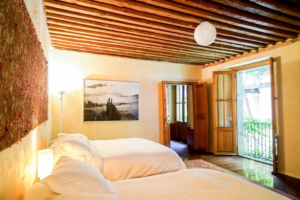 A bedroom with two beds, a wooden ceiling, a painting on the wall, and a view of the outside balcony.