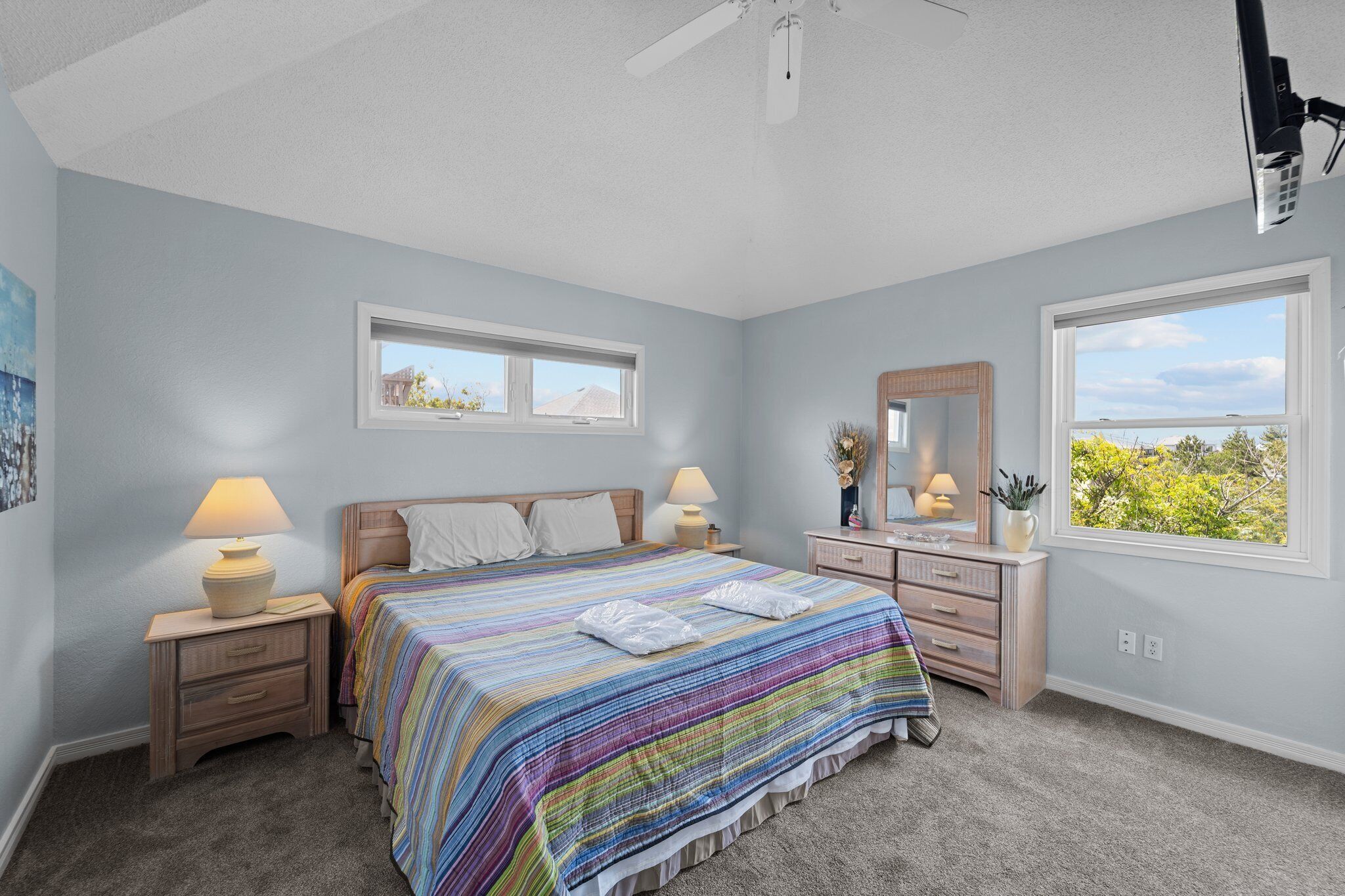 A bedroom with a large bed, two bedside tables with lamps, a dresser, a mirror, and two windows offering a view of greenery.