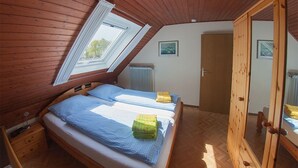 Una habitación con techo inclinado, paredes de madera, una cama con ropa de cama azul, un armario de madera y una ventana con vistas a árboles.