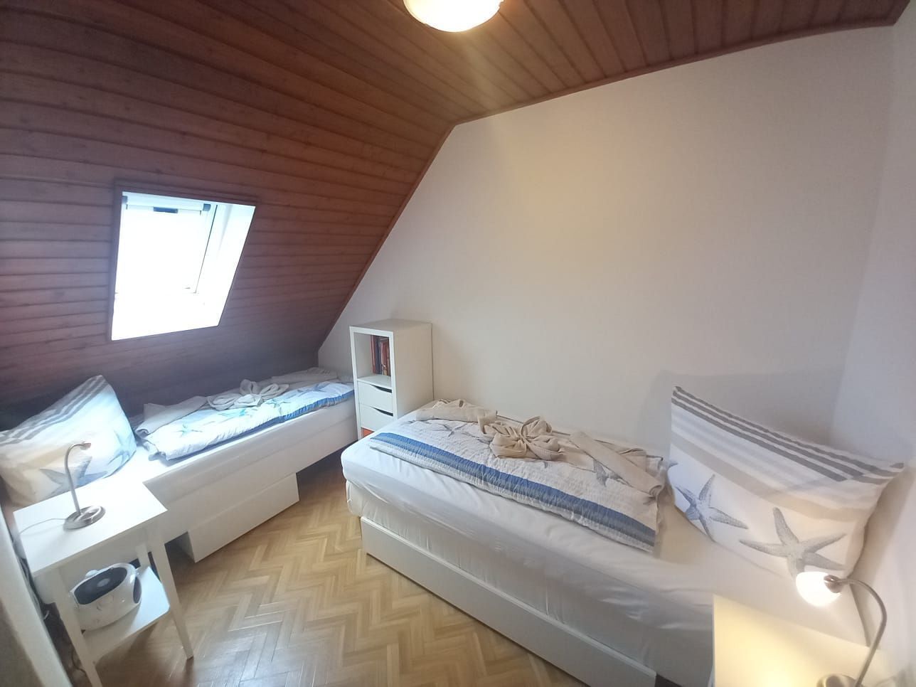 Ett litet, ombonat rum med två enkelsängar, ett vitt skrivbord med en lampa och ett litet vitt skåp.