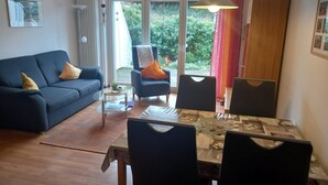 Una sala de estar moderna con un sofá azul, una mesa de comedor con sillas y un gran ventanal con vista a la vegetación.