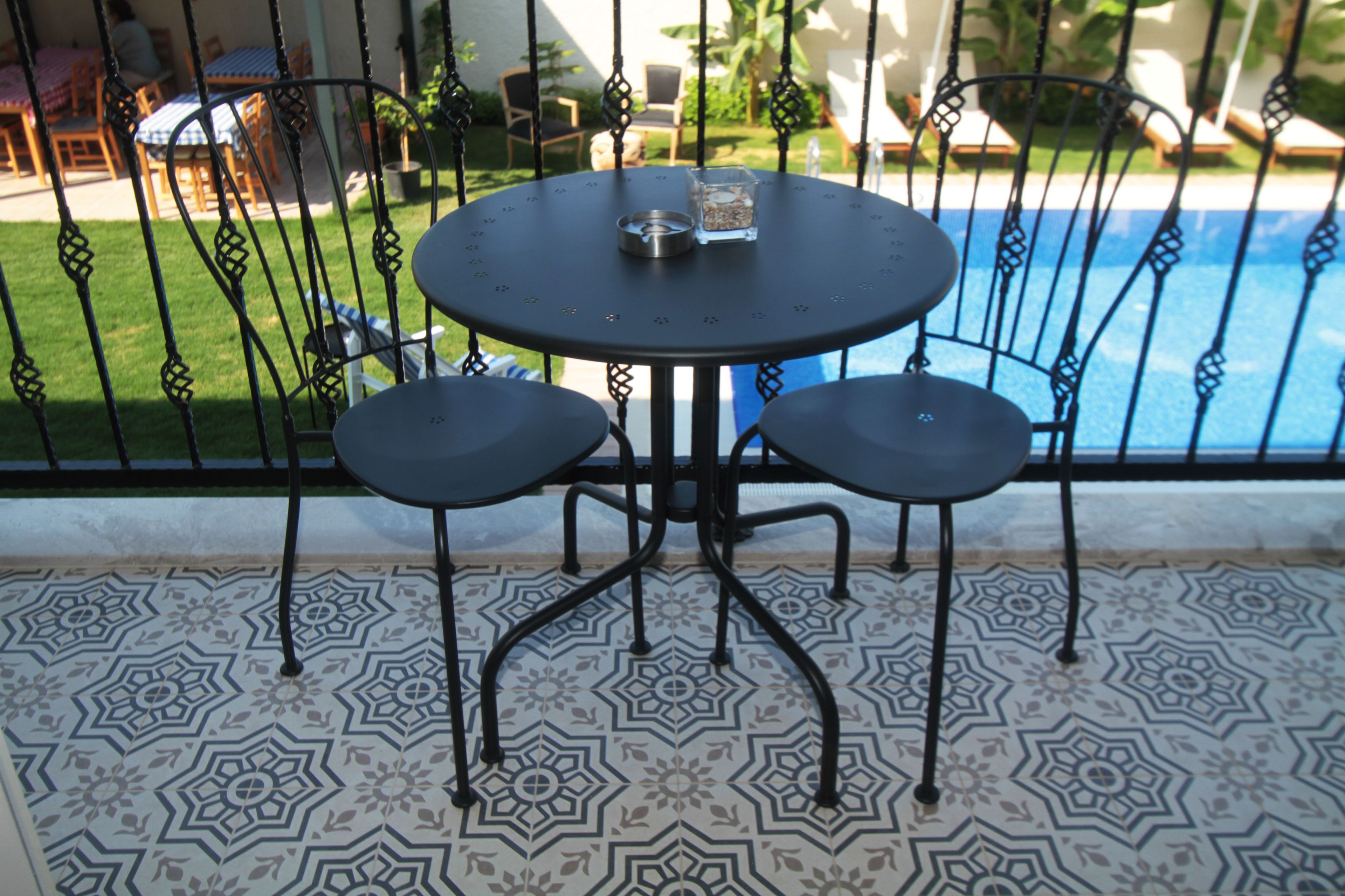 A round table with two chairs, a glass, and a metal tray on a patterned floor, overlooking a swimming pool.