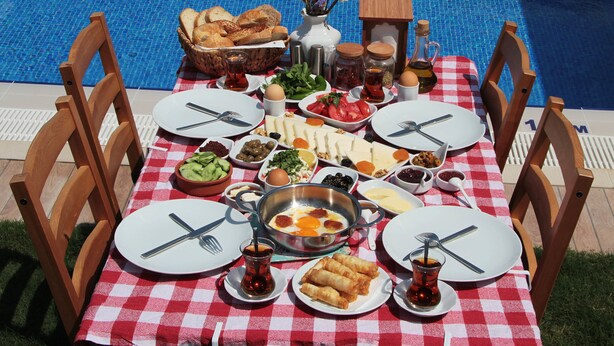 A dining table with a checkered tablecloth, surrounded by wooden chairs, set with plates, cutlery, and various dishes, including eggs and bread, by a pool. - Duman Boutique Hotel (Cesme)