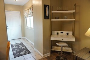 Um corredor com uma porta branca, uma janela com cortinas florais, uma escrivaninha branca com uma cadeira e uma prateleira com objetos decorativos.