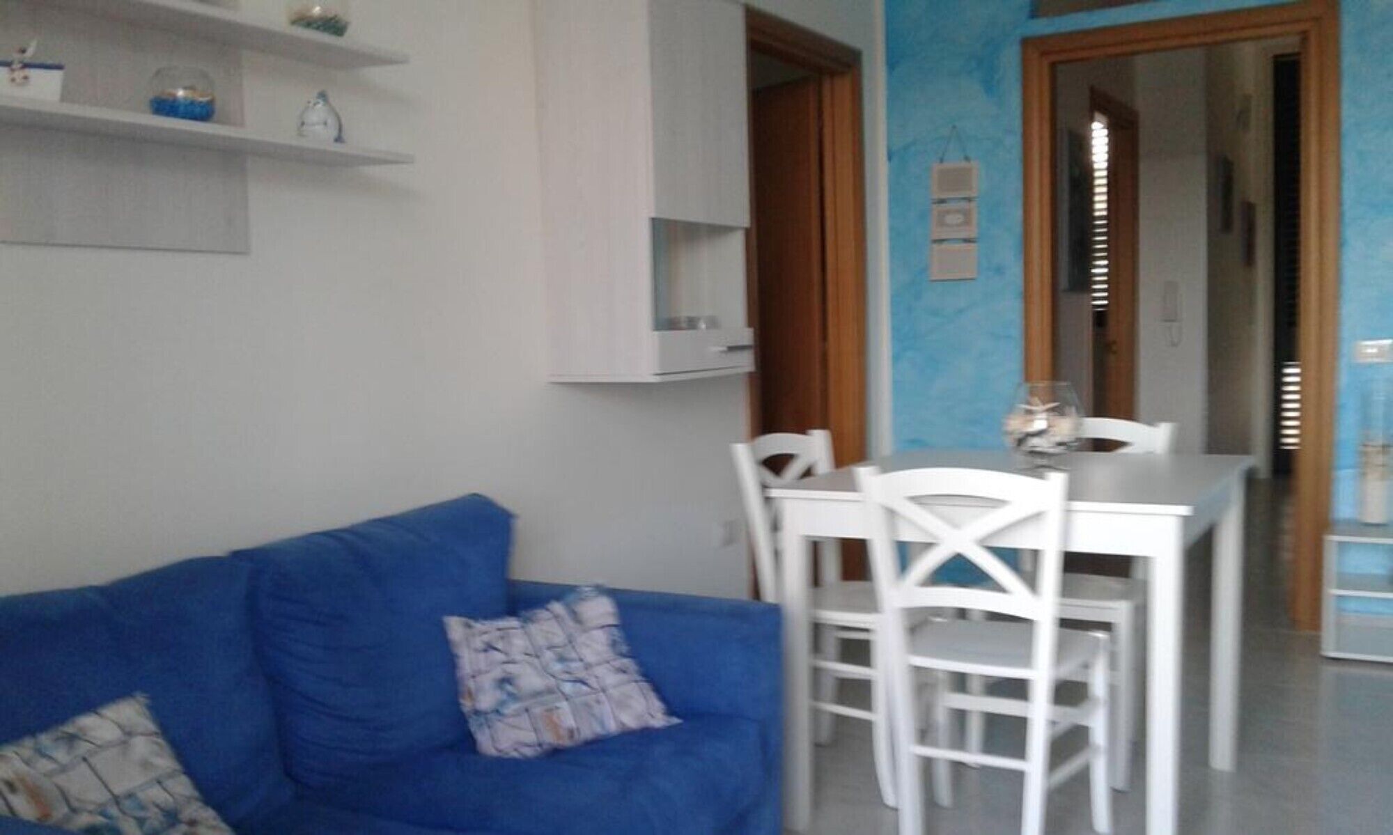 Una sala de estar con un sofá azul, sillas blancas, una mesita y una puerta que da a un comedor.