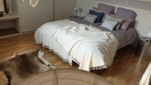 Quarto com cama, travesseiros, manta branca, tapete de pele, luminária de cabeceira e lixeira.