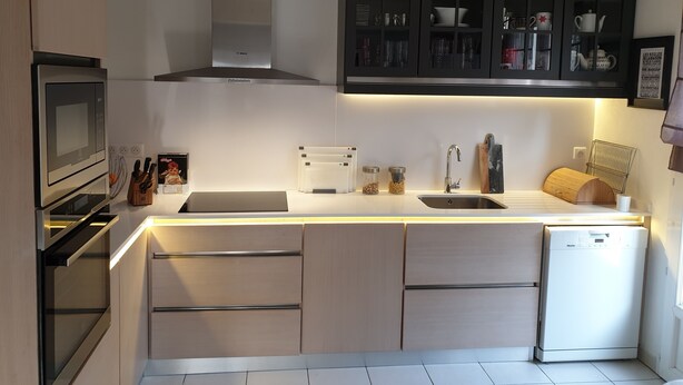Uma cozinha moderna com forno embutido, micro-ondas e máquina de lavar loiça. Possui uma ilha grande com pia e uma luminária suspensa.