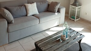 Uma sala de estar com um sofá cinzento, uma mesa de centro de madeira com objetos decorativos, uma parede branca com um quadro e um candeeiro branco.