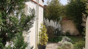 Um jardim com caminho de pedra, diversas plantas e um edifício com fachada branca e telhado vermelho.