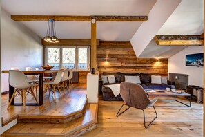 A modern interior with a wooden staircase, a dining area with chairs and a table, a sofa set, and a television mounted on the wall.