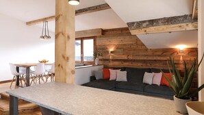 Een moderne keuken met een houten accentmuur, een eetgedeelte met witte stoelen en een grijze bank met kleurrijke kussens.