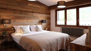 Een slaapkamer met een houten wand, een bed met witte beddengoed, twee bedlampjes en een raam met uitzicht op bomen.
