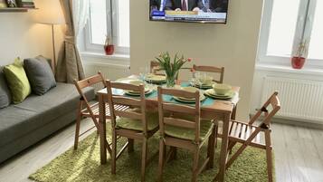 Uma sala de jantar com mesa posta para quatro pessoas, cadeiras, televisão fixada na parede e uma janela com cortinas.