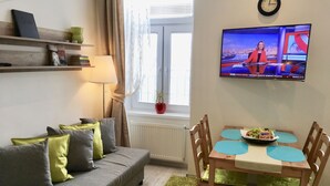A living room with a grey sofa, a wooden dining table with chairs, a TV on the wall, and a window with curtains. - Johansson Apartments Vienna (Vienna)
