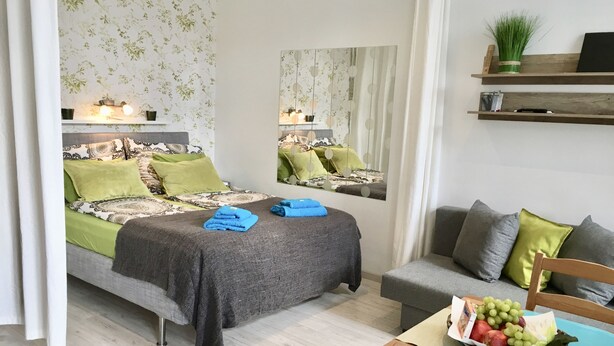 A well-lit bedroom with a bed, bedside table, and a mirror. There is a grey sofa and a wooden chair in the room. - Johansson Apartments Vienna (Vienna)