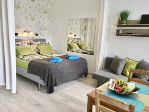 A well-lit bedroom with a bed, bedside table, and a mirror. There is a grey sofa and a wooden chair in the room. - Johansson Apartments Vienna (Vienna)