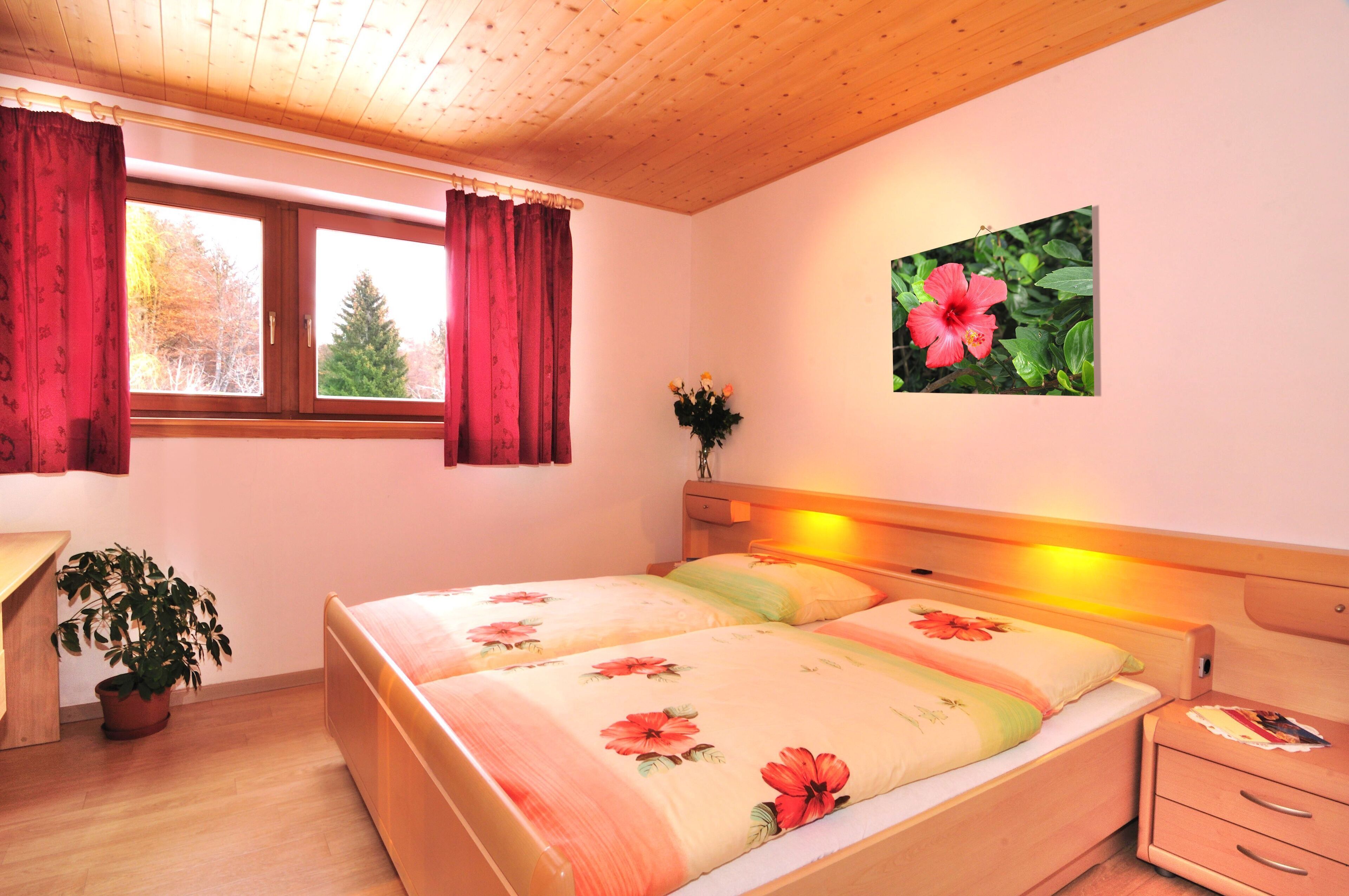 A bedroom with two beds, a wooden ceiling, a window with red curtains, a wall-mounted picture of a flower, and a bedside table with a lamp.
