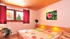 Ett sovrum med två sängar, ett träpanelat tak, ett fönster med röda gardiner, en tavla med ett blommotiv som hänger på väggen och ett nattduksbord med en lampa.