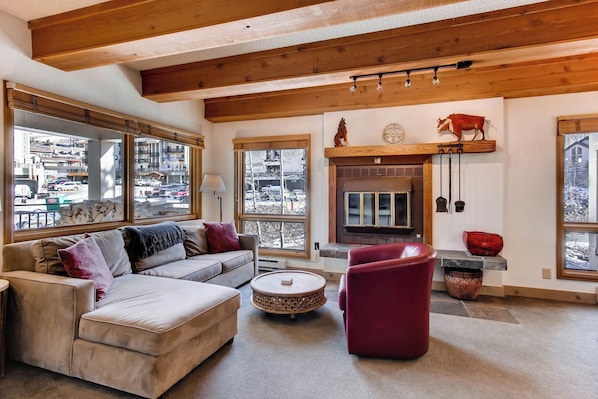 A cozy living room with a fireplace, large windows, and a wooden ceiling.
