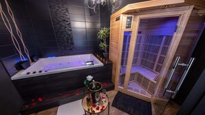 Moderni kylpyhuone, jossa on suuri kylpyamme, sauna ja pieni pöytä, jonka päällä on maljakko ja laseja.
