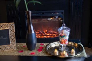 Een open haard met brandhout, een vaas, een dienblad met kaarsen, en een decoratief object op een tafel.