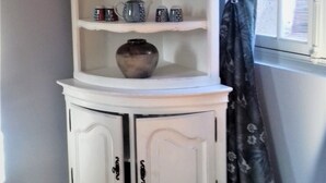 A white corner cabinet with shelves and doors, placed against a wall with a window and curtains. - cottage of the Small Mass (Amboise)