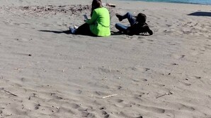 Una spiaggia sabbiosa con due persone sedute, un mare blu limpido e edifici sullo sfondo.