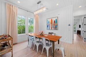 A dining area with a wooden table, white chairs, a chandelier, and a large window with curtains.