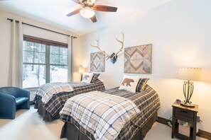 A bedroom with two beds, a ceiling fan, a window with curtains, and wall decorations including animal antlers and geometric patterns.