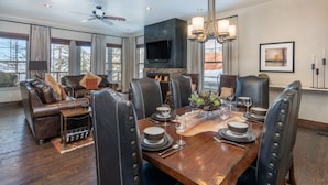 Comedor con mesa de madera dispuesta para seis comensales, sillas de cuero, chimenea y televisor montado en la pared.