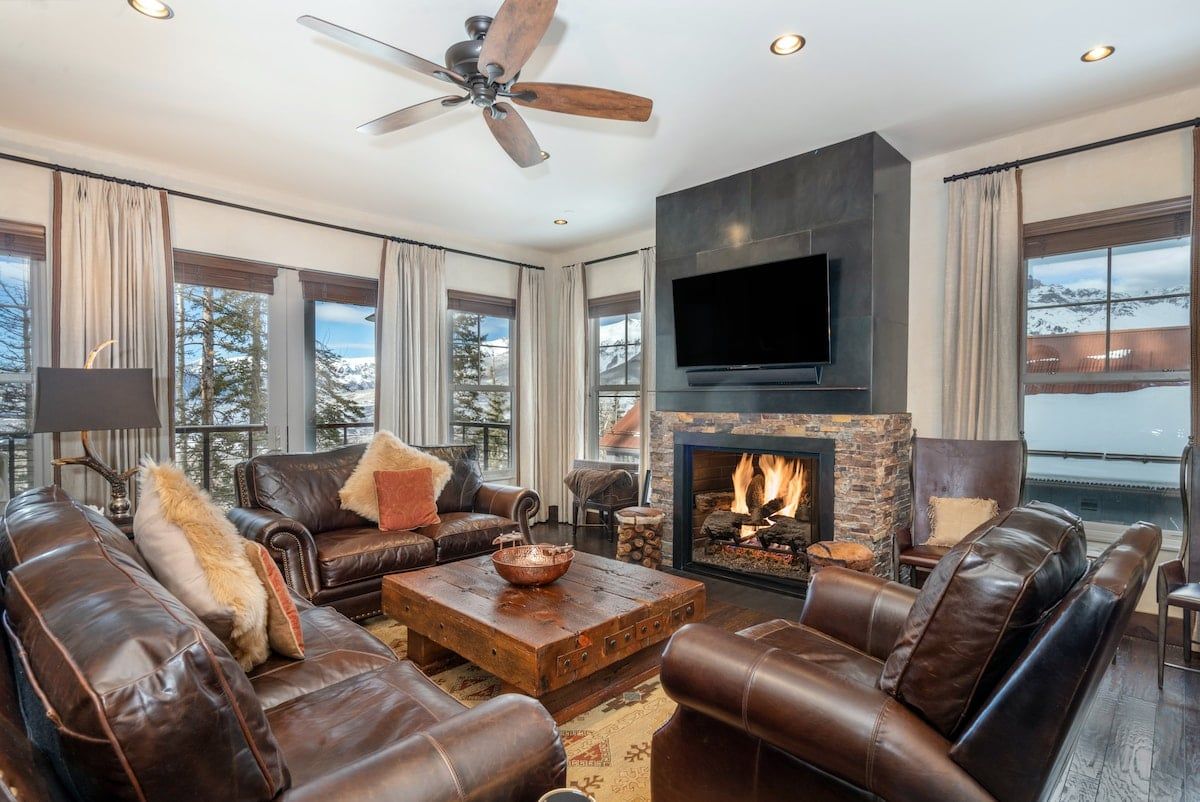 A modern living room with leather sofas, a fireplace, a flat-screen TV, and a ceiling fan.
