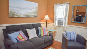 A living room with a grey sofa, a small armchair, a lamp, a window with curtains, and a painting on the wall.