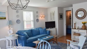 A living room with a blue sofa, a television, a small table, and a dining area with chairs and a table. - End Unit Condo with breathtaking ocean views! (Dennis Port)