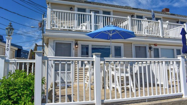 A two-story building with white railings and a blue umbrella. - End Unit Condo with breathtaking ocean views! (Dennis Port)