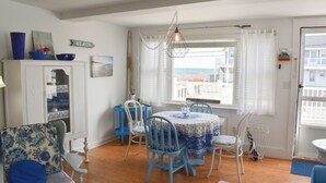 A dining area with a table and chairs, a view of the sea through a window, and a sign indicating a beach. - End Unit Condo with breathtaking ocean views! (Dennis Port)