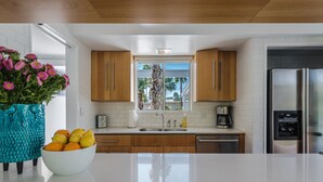 Una cocina moderna con armarios de madera, encimera blanca, refrigerador de acero inoxidable y ventana con vista a palmeras.