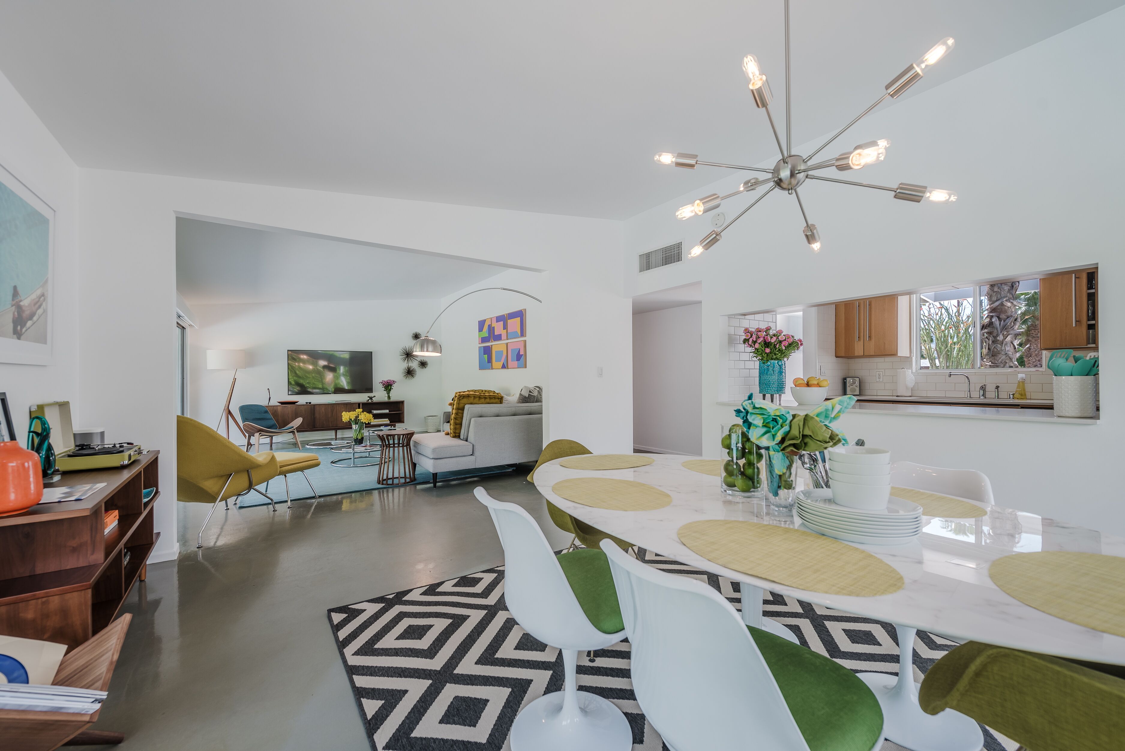 A modern living room with a dining area, featuring a large round table, white and green chairs, a chandelier, and a television in the background.