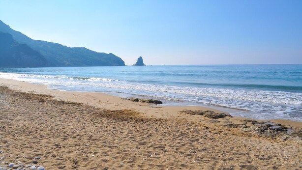 Une plage avec un ciel dégagé, une eau calme et un promontoire rocheux à l’horizon.