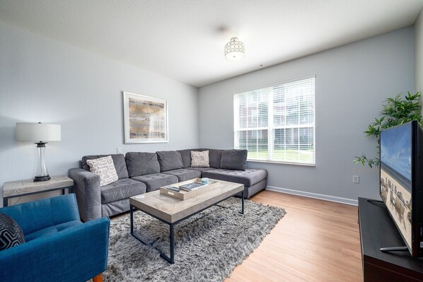 Una sala de estar moderna con un sofá gris, un sillón azul, una mesita de centro, una lámpara y un amplio ventanal con persianas.