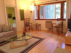 Una sala de estar con una mesa de centro de vidrio, un sofá, dos sillas de madera, una mesita auxiliar y un radiador.