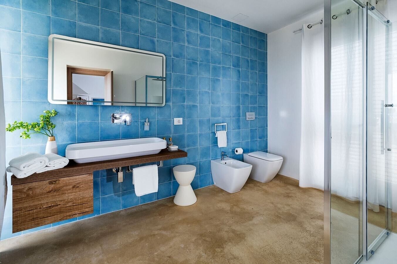 A modern bathroom with blue tiles, a wooden vanity, a white sink, a bidet, and a glass shower enclosure.