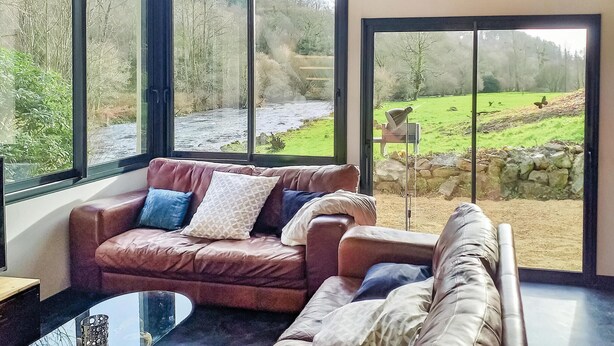 Een moderne woonkamer met een leren bank, een glazen salontafel en grote ramen met uitzicht op een rivier en groen landschap.