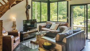 Een knusse woonkamer met een televisie, leren banken en een groot raam met uitzicht op groen.