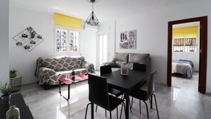A modern living room with a sofa, dining table, and chairs. There is a window with a view of the outside, and a door leading to another room. - 1028 Casa Evelyn D (Nerja)
