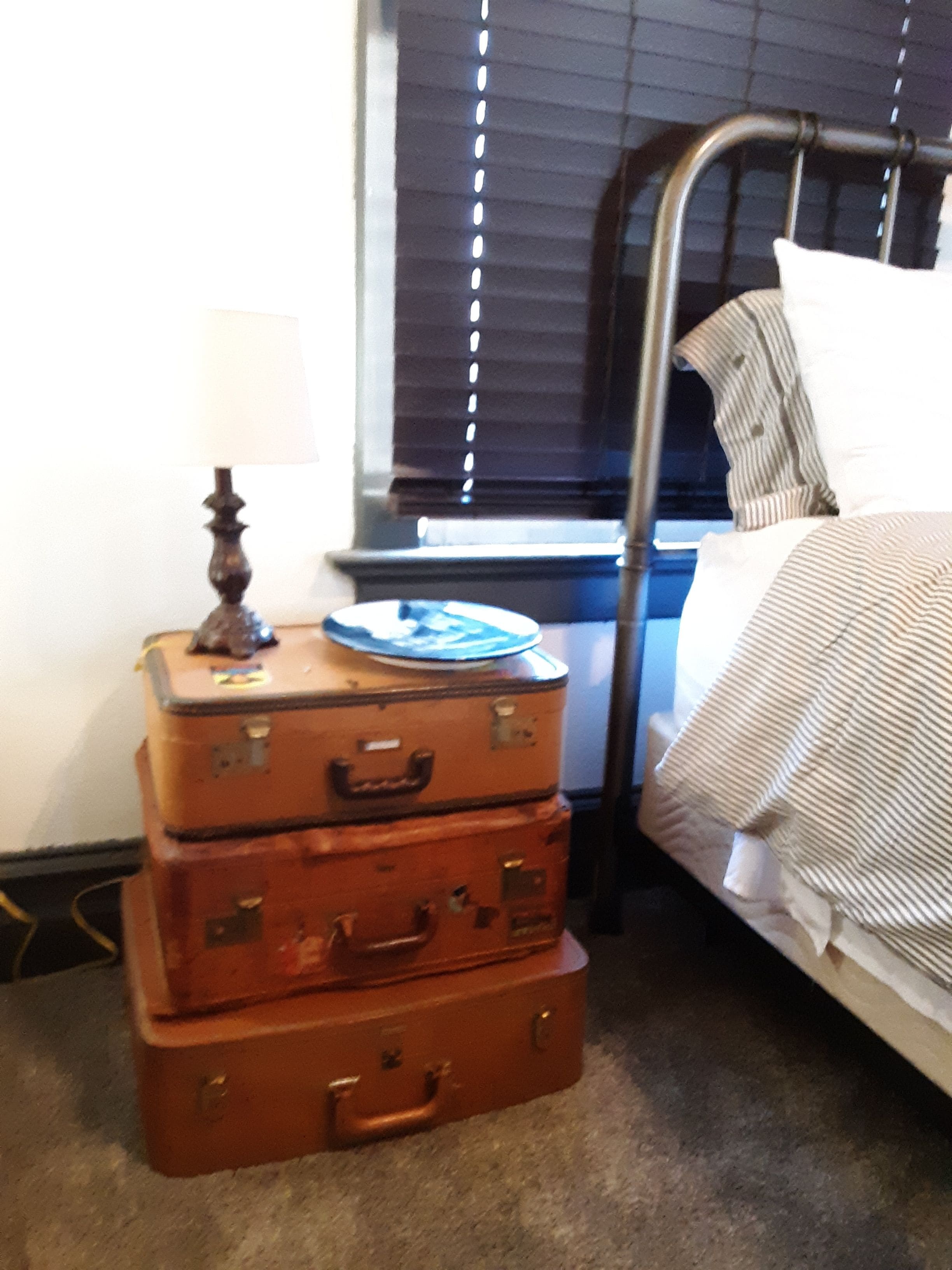 A bed with striped bedding, a metal headboard, and a window with blinds. There is a stack of three brown suitcases with a plate on top, and a lamp on the floor beside the suitcases.