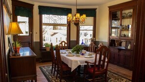 A dining room with a wooden table set for four, surrounded by chairs. There is a chandelier above the table, a cabinet with dishes to the right, and a window with curtains. - The Katherine Holle House (Watertown)