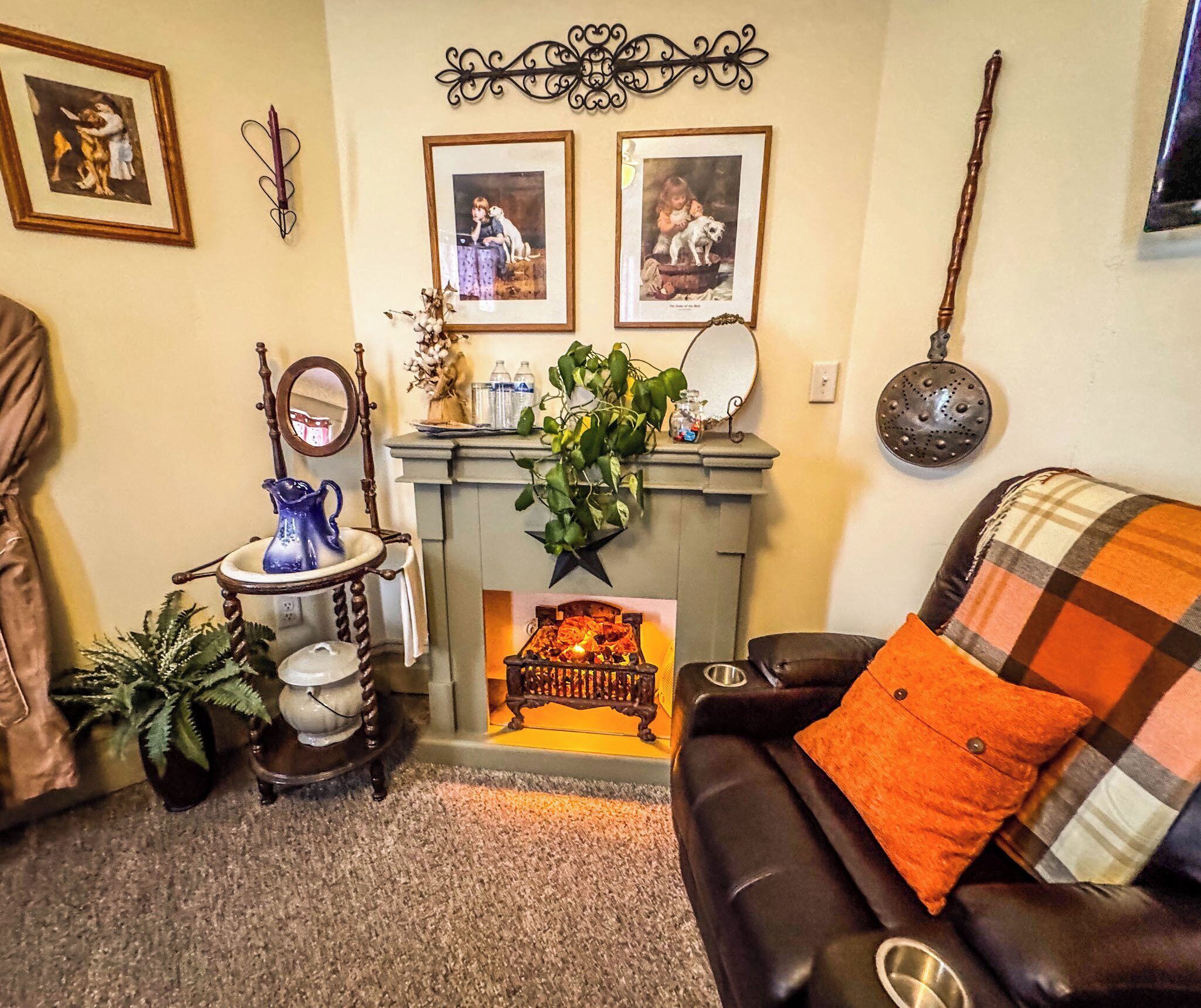 A cozy living room with a fireplace, a black leather sofa with an orange pillow, a wooden side table with a blue pitcher, and framed pictures on the wall.