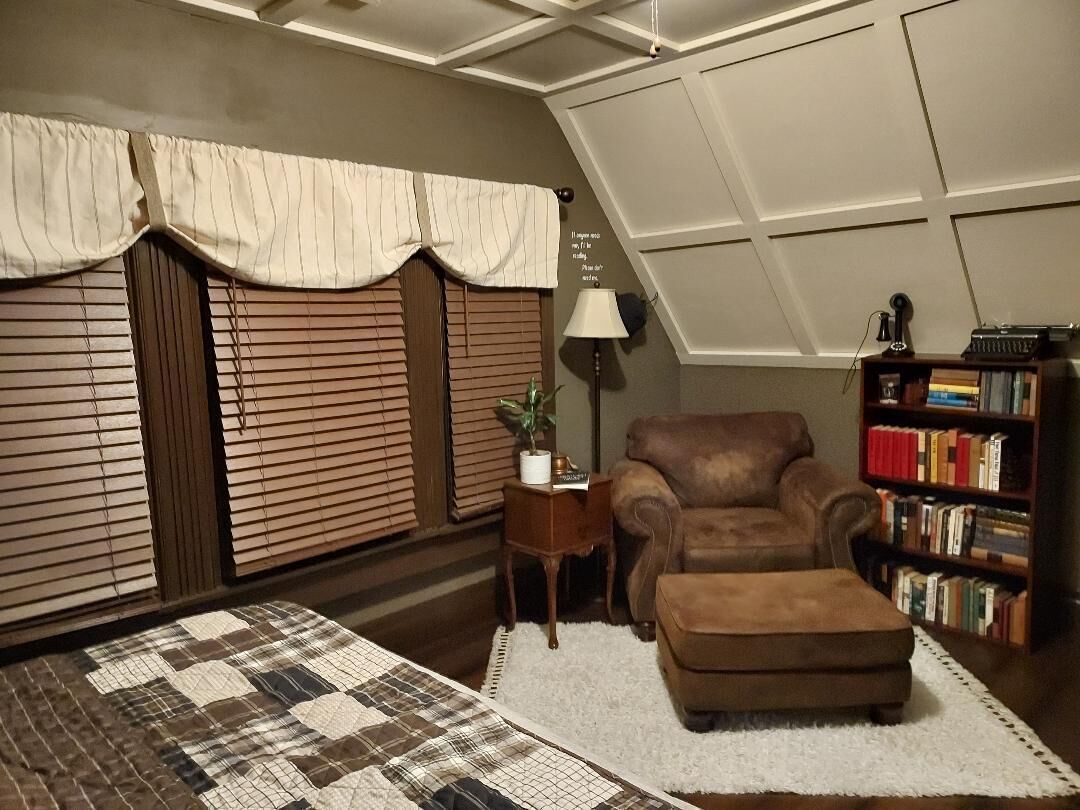 Un cuarto acogedor con un sillón de cuero marrón, una mesita de noche con una lámpara, una estantería y un colcha en el suelo.