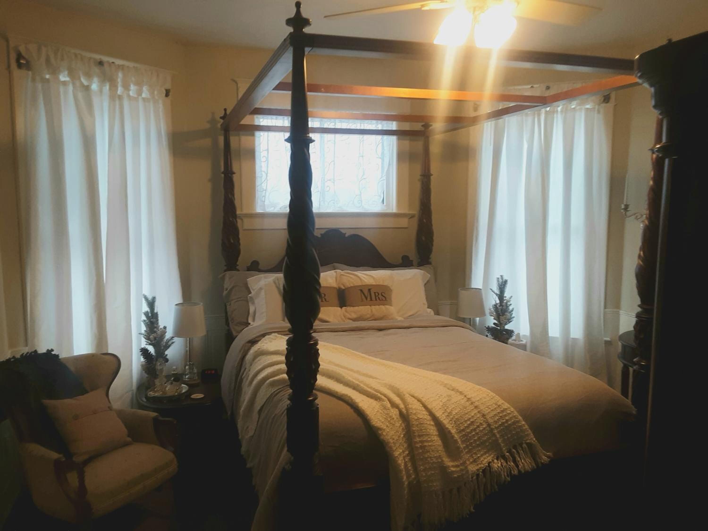 A four-poster canopy bed with a 'Mrs' pillow, a chair, a nightstand with a lamp, and a small Christmas tree.