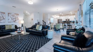 A modern living room with a black leather sofa, a glass coffee table, and a patterned rug. There is a dining area with a table and chairs, and a kitchen in the background. - Modern 7BR w/Pool/ Minutes from Disney/ ESPN (Kissimmee)